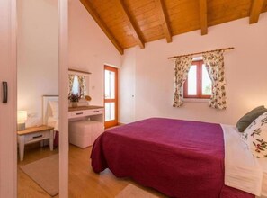 A bedroom with a wooden ceiling, a bed with a red blanket, a dresser, a mirror, and a window with floral curtains. - The Grandfather Villa Heated Pool Retreat (Stankovci)