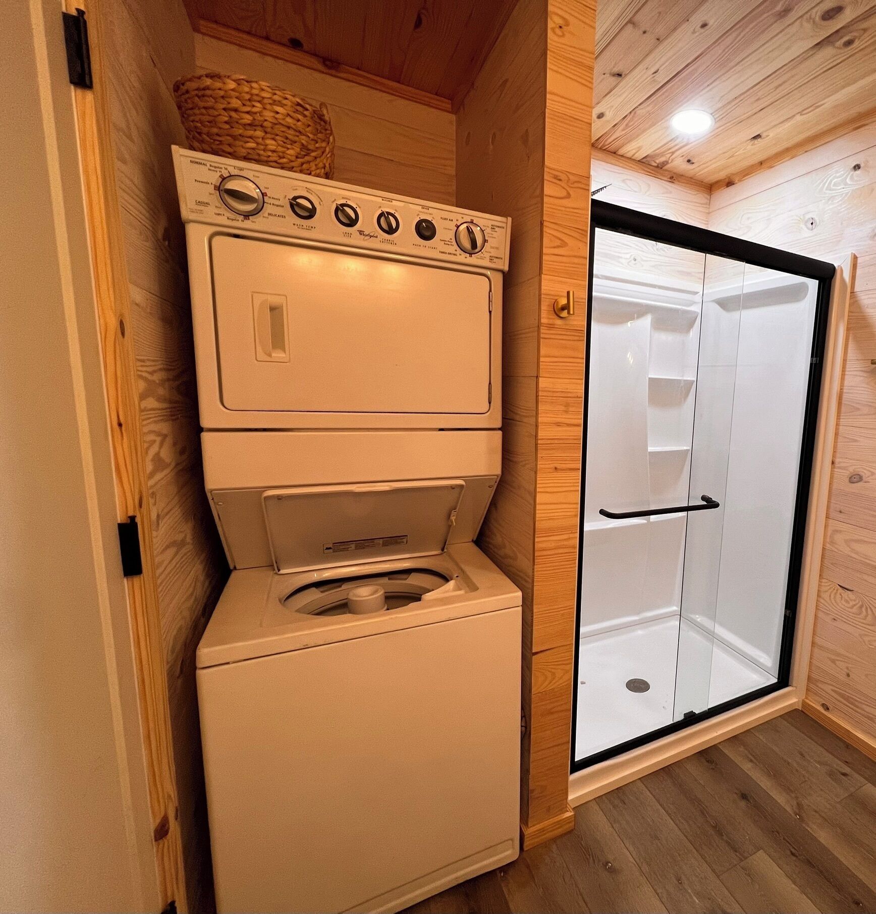 A compact laundry room with a stacked washer and dryer, a wooden ceiling, and a white shower enclosure.
