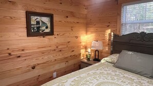 A wooden-paneled room with a bed, a nightstand with a lamp, and a framed picture on the wall. - Peaceful 2BR cabin between Montgomery & Troy, AL with trails and stocked pond (Ramer)