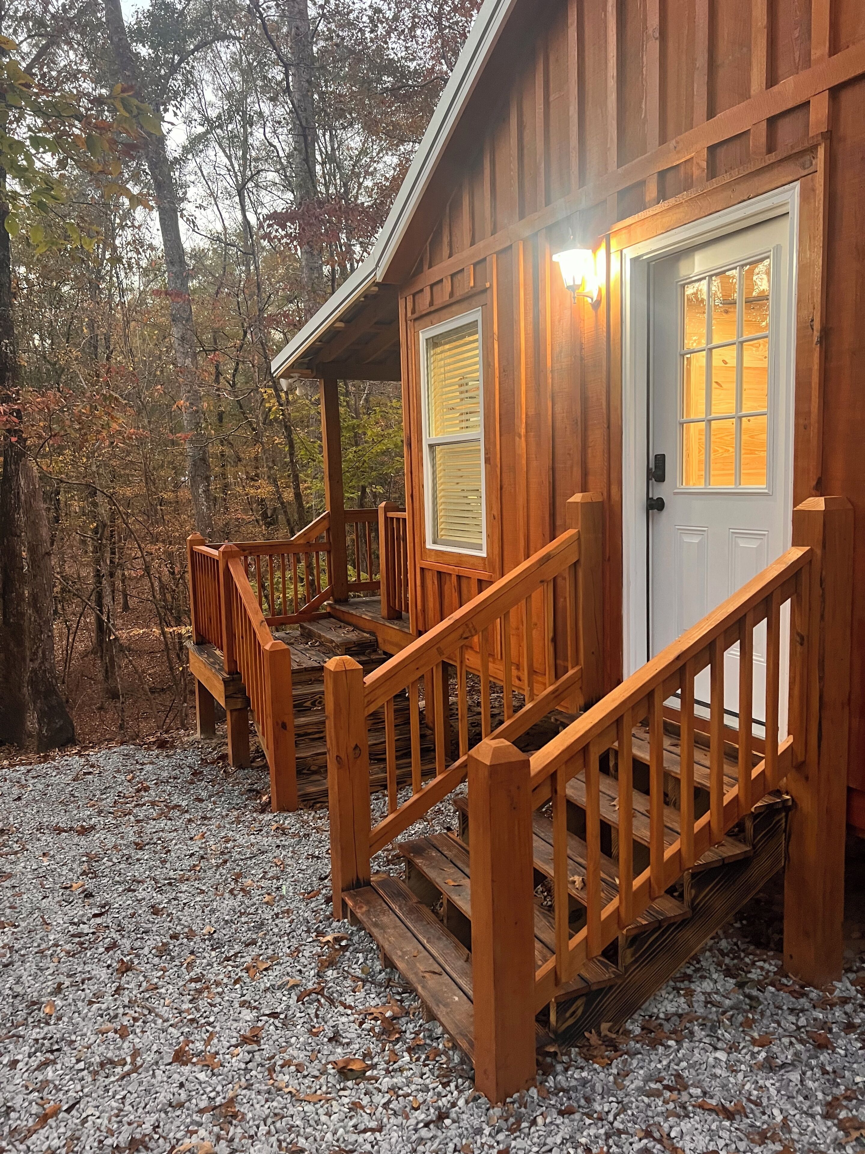 A wooden cabin with a porch and a door, surrounded by trees and gravel.