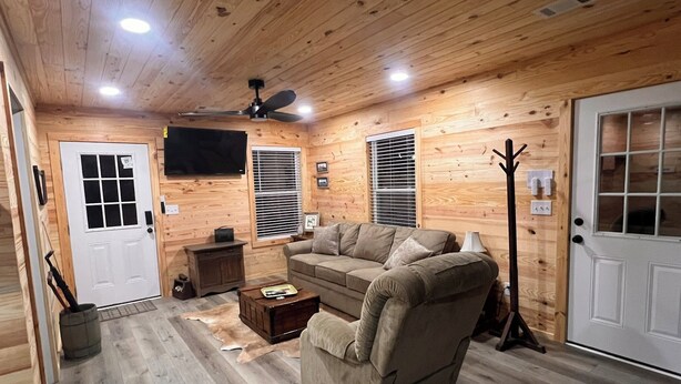 A cozy cabin interior with a wooden ceiling, a ceiling fan, a television mounted on the wall, a sofa, a coffee table, and a door with a window. - Peaceful 2BR cabin between Montgomery & Troy, AL with trails and stocked pond (Ramer)