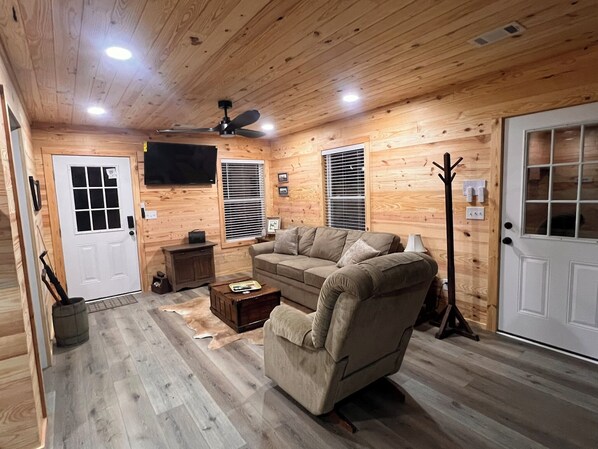 A cozy cabin interior with a wooden ceiling, a ceiling fan, a television mounted on the wall, a sofa, a coffee table, and a door with a window.