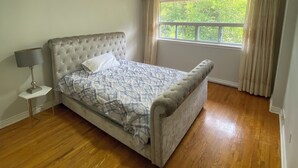 A bedroom with a tufted headboard bed, a bedside lamp, a window with curtains, and a wooden floor. - Cozy Home in North York Toronto (Toronto)