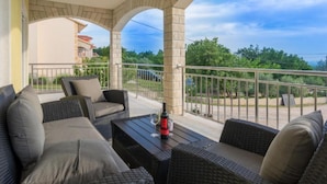 A covered outdoor seating area with wicker furniture and a table, overlooking a green landscape and a building in the background. - Holiday House Balen With Swimming Pool (Benkovac)