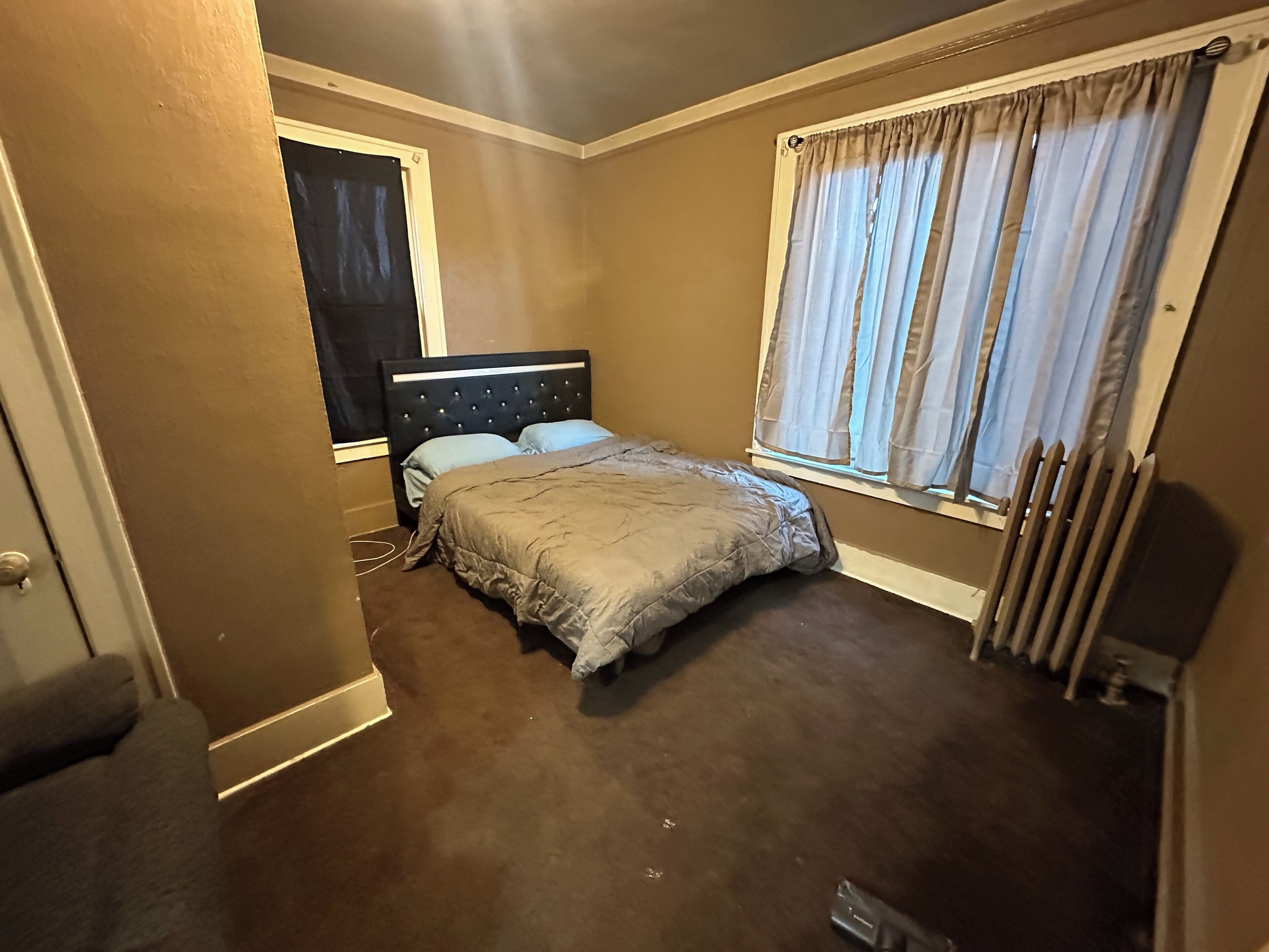 A bedroom with a bed, a window with curtains, a radiator, and a door.
