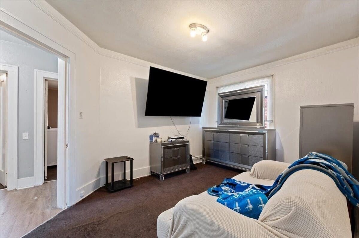 A room with a flat-screen TV mounted on the wall, a small cabinet, a bed with a blue blanket, and a window above the cabinet.