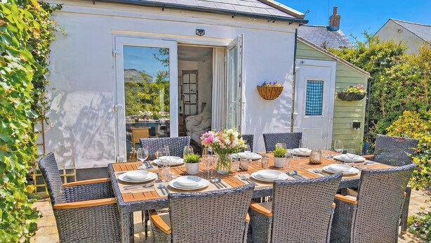 An outdoor dining area with wicker chairs and a table set for six, a white exterior wall, and a door leading to an interior space. - 4 bedroom accommodation in California (California)