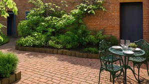 A brick patio with a round table and chairs, surrounded by lush greenery and a brick wall. - The Coach House - UK50884 (North Walsham)