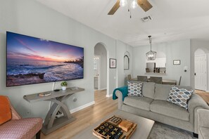 Un salon doté d’un grand téléviseur à écran plat, d’un canapé gris, d’une table basse sur laquelle sont posés des jeux de société et d’un ventilateur de plafond.