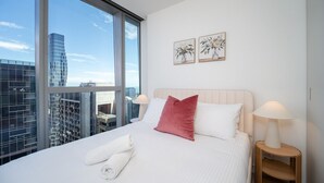 A hotel room with a large window, a bed with white bedding, a red pillow, two bedside lamps, and two framed floral paintings on the wall. - West Side Place Family Suite w/ Pool+Sauna+Gym (Melbourne)