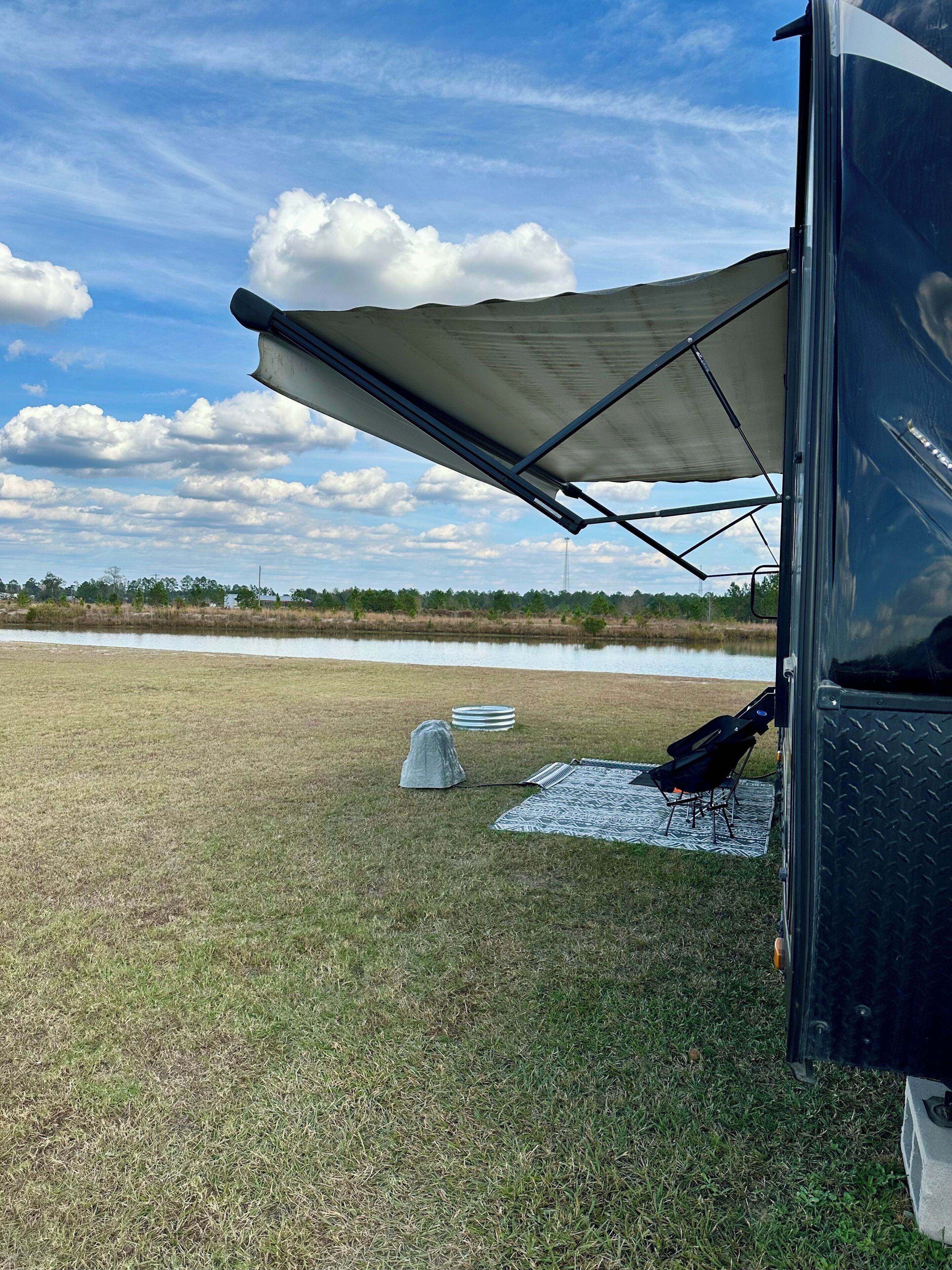 A camping setup with a tent, a portable grill, and a picnic table in a grassy field with a water body in the background.