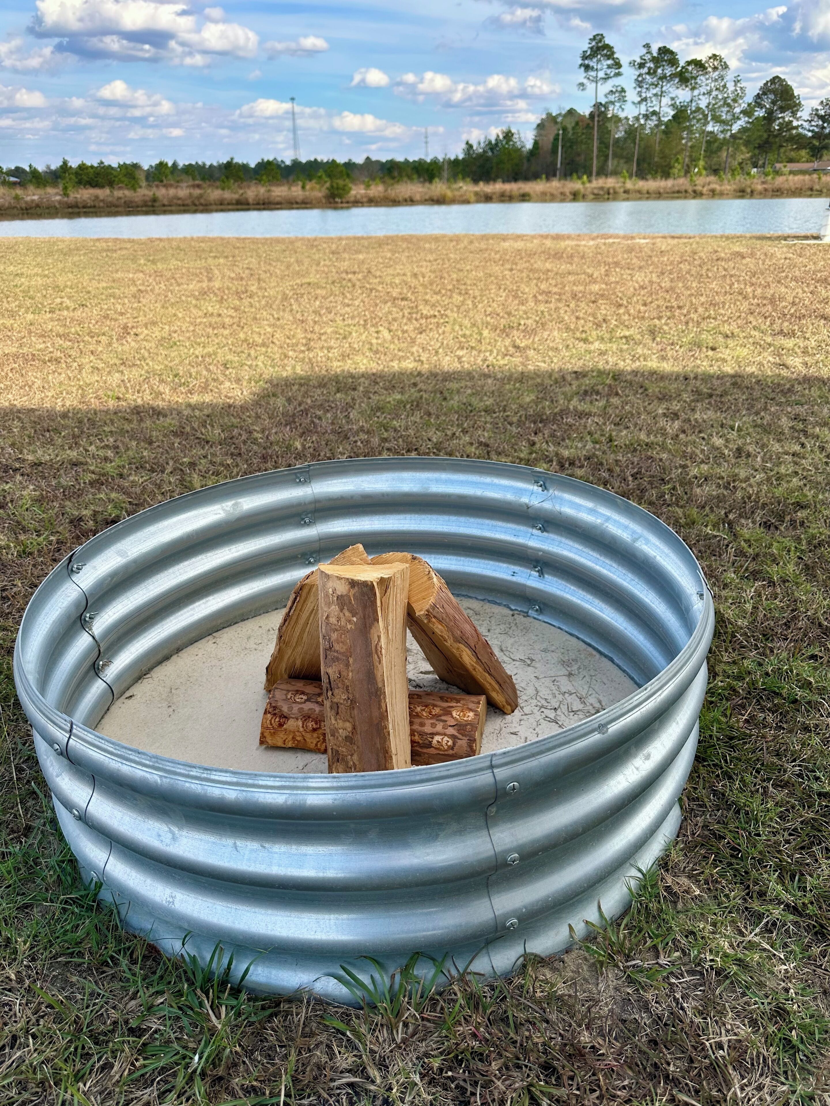 A metal fire pit with logs and sand, surrounded by grass and a body of water in the background.