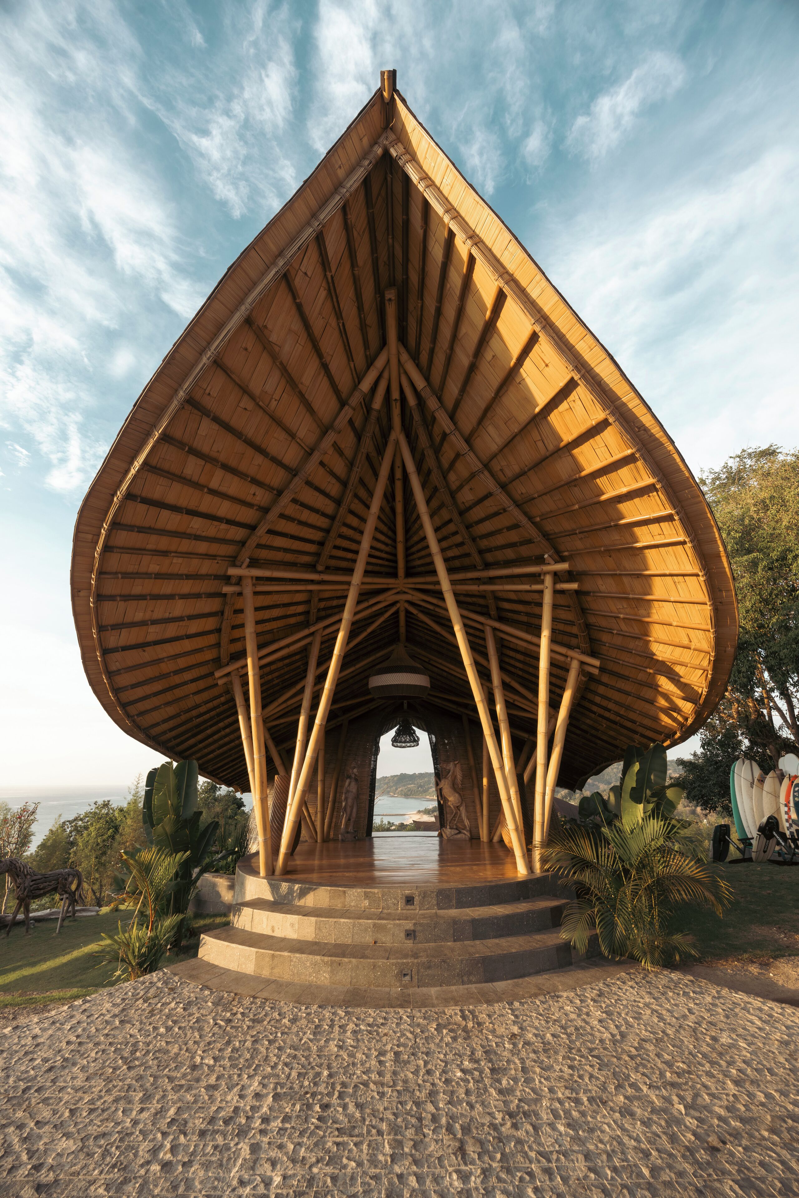 A large, open pavilion with a triangular roof structure, surrounded by a stone pathway and palm trees.