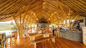 Un comedor con techo de bambú, mesas y sillas de madera, una barra y vistas al paisaje.