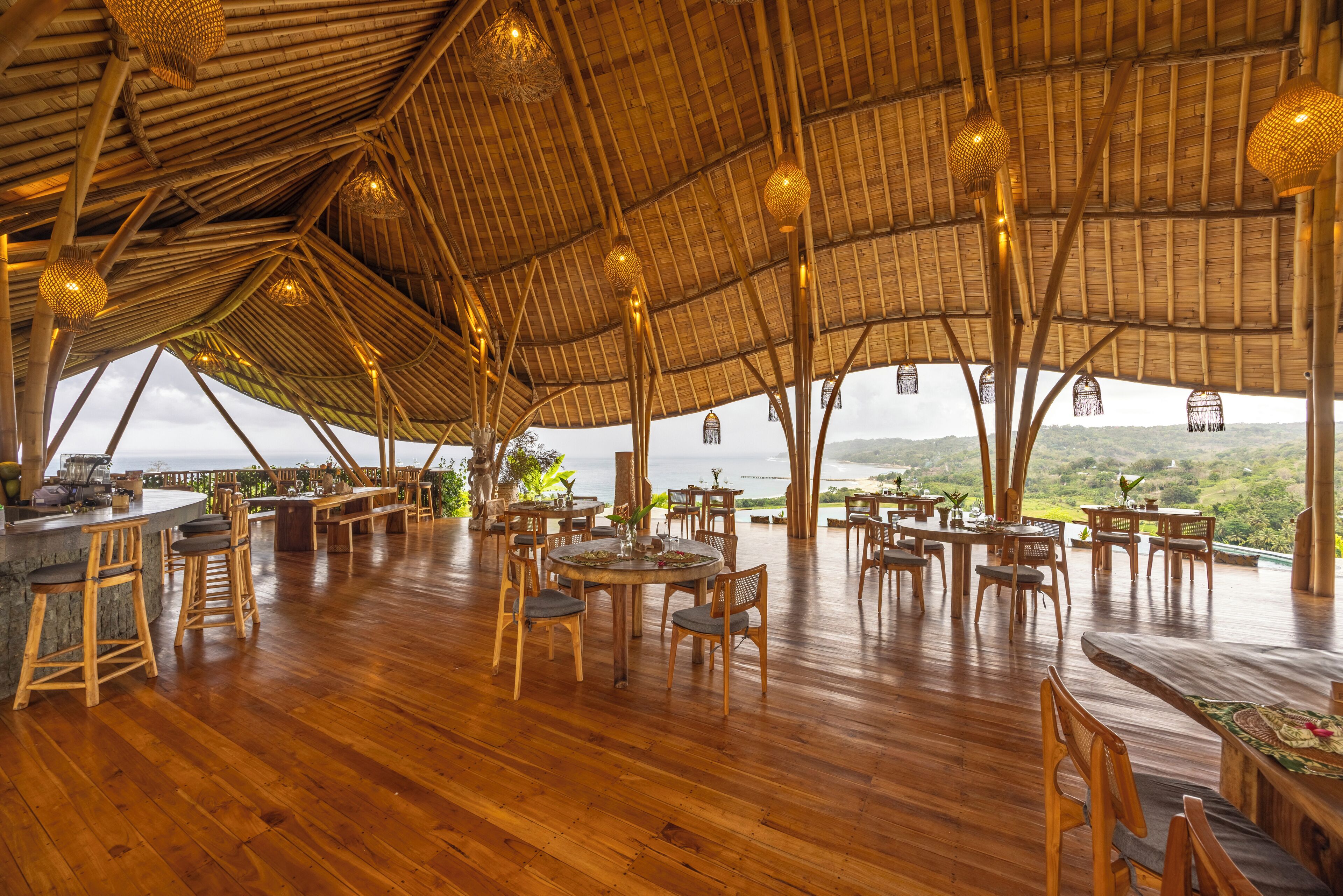 A spacious dining area with wooden tables and chairs, a bar area, and a thatched roof structure providing a view of the landscape.