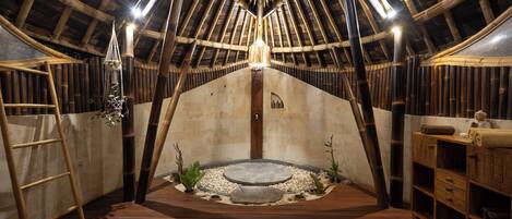 An attic room with a sloped ceiling, wooden beams, a central light fixture, a small circular stone feature, and a wooden ladder.