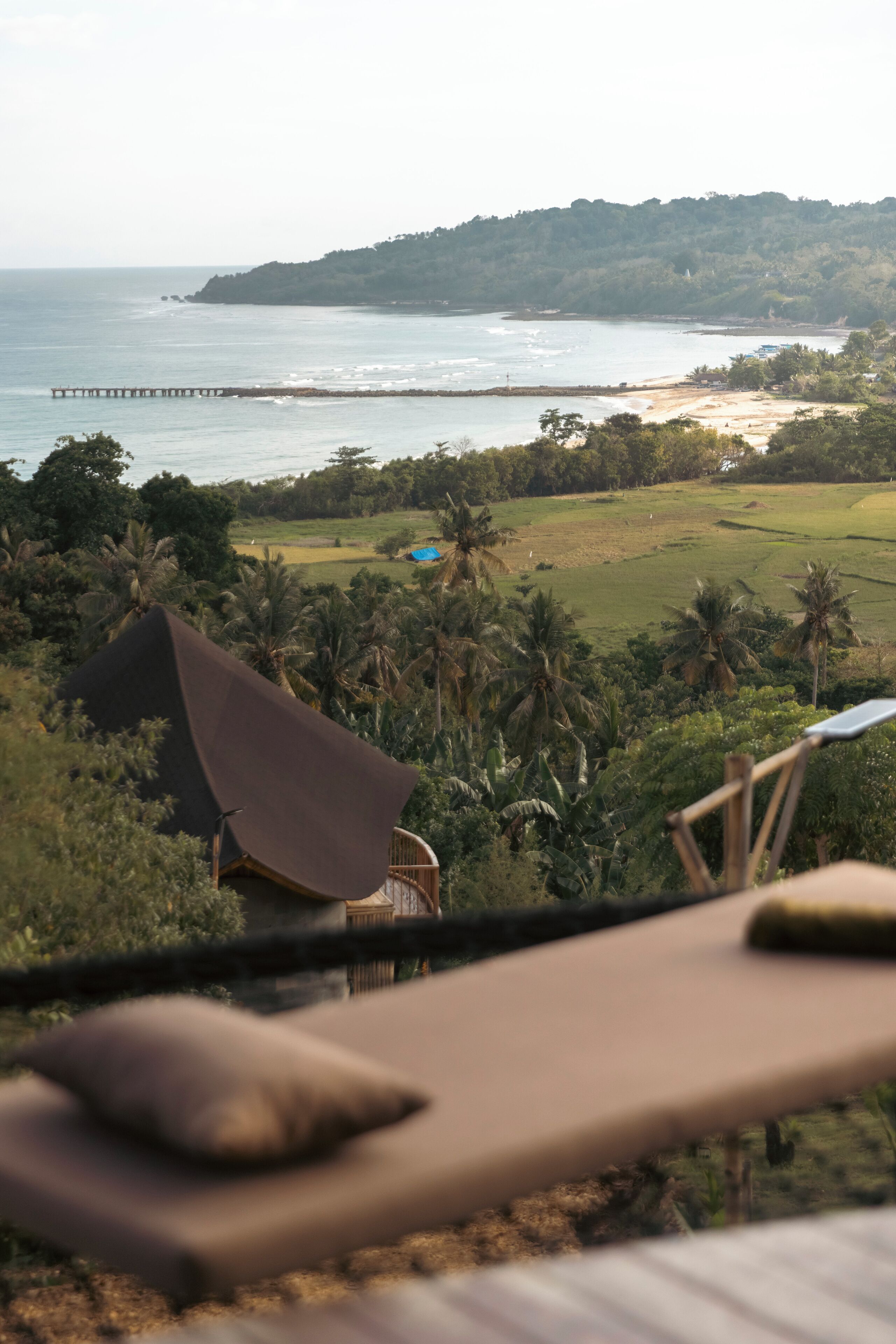 A view from a balcony overlooking a beach with a pier, palm trees, and a distant hill.