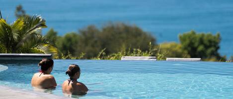 A swimming pool with two people in the water, surrounded by greenery and a view of the ocean.