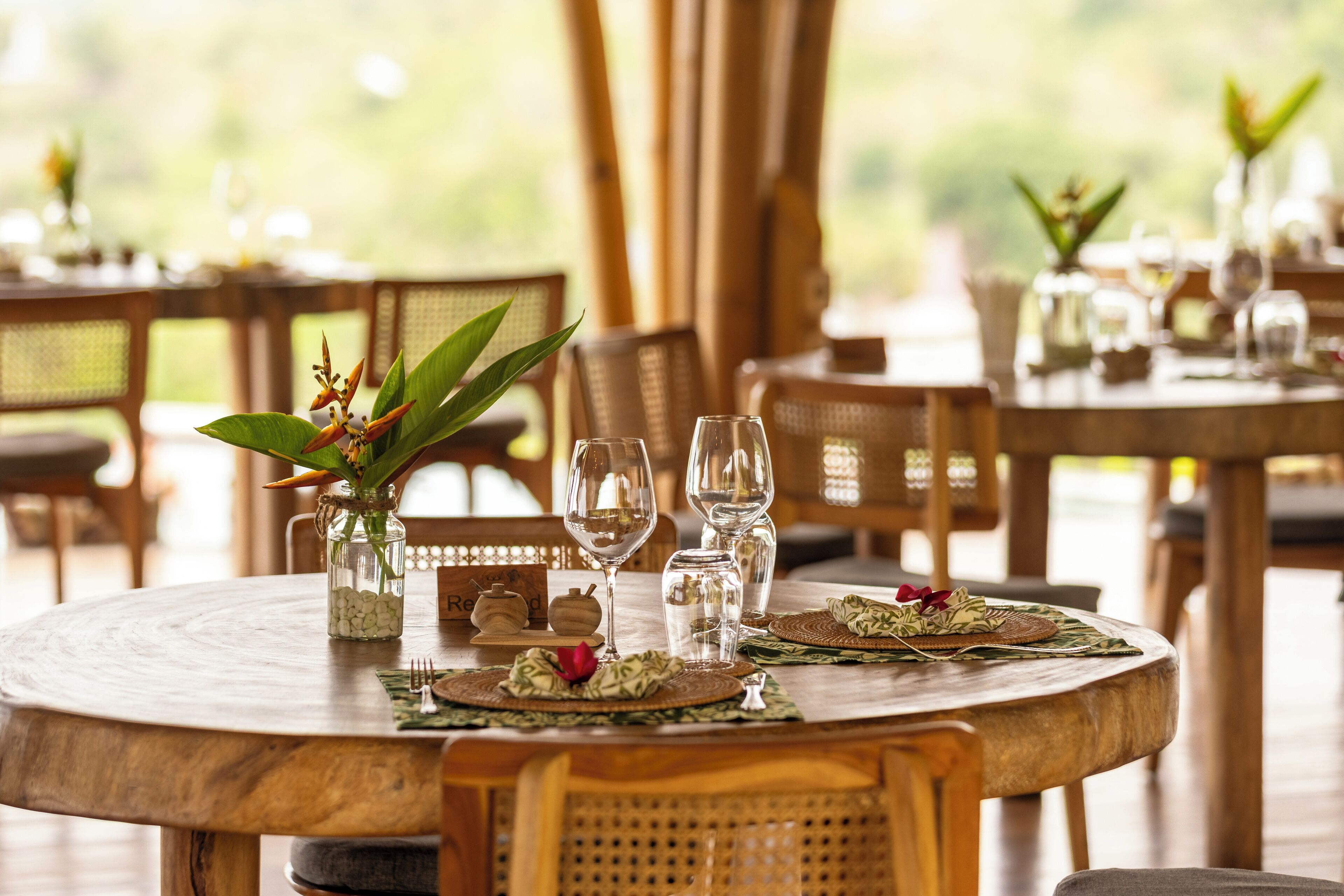 A round wooden dining table set with glassware, woven placemats, and a centerpiece of green leaves and a red flower.