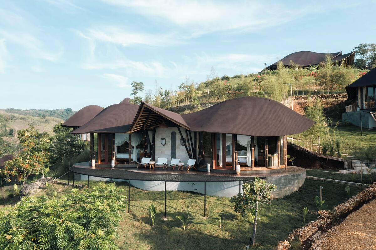 A modern villa with a curved roof, a covered outdoor seating area, and a view of the surrounding landscape.