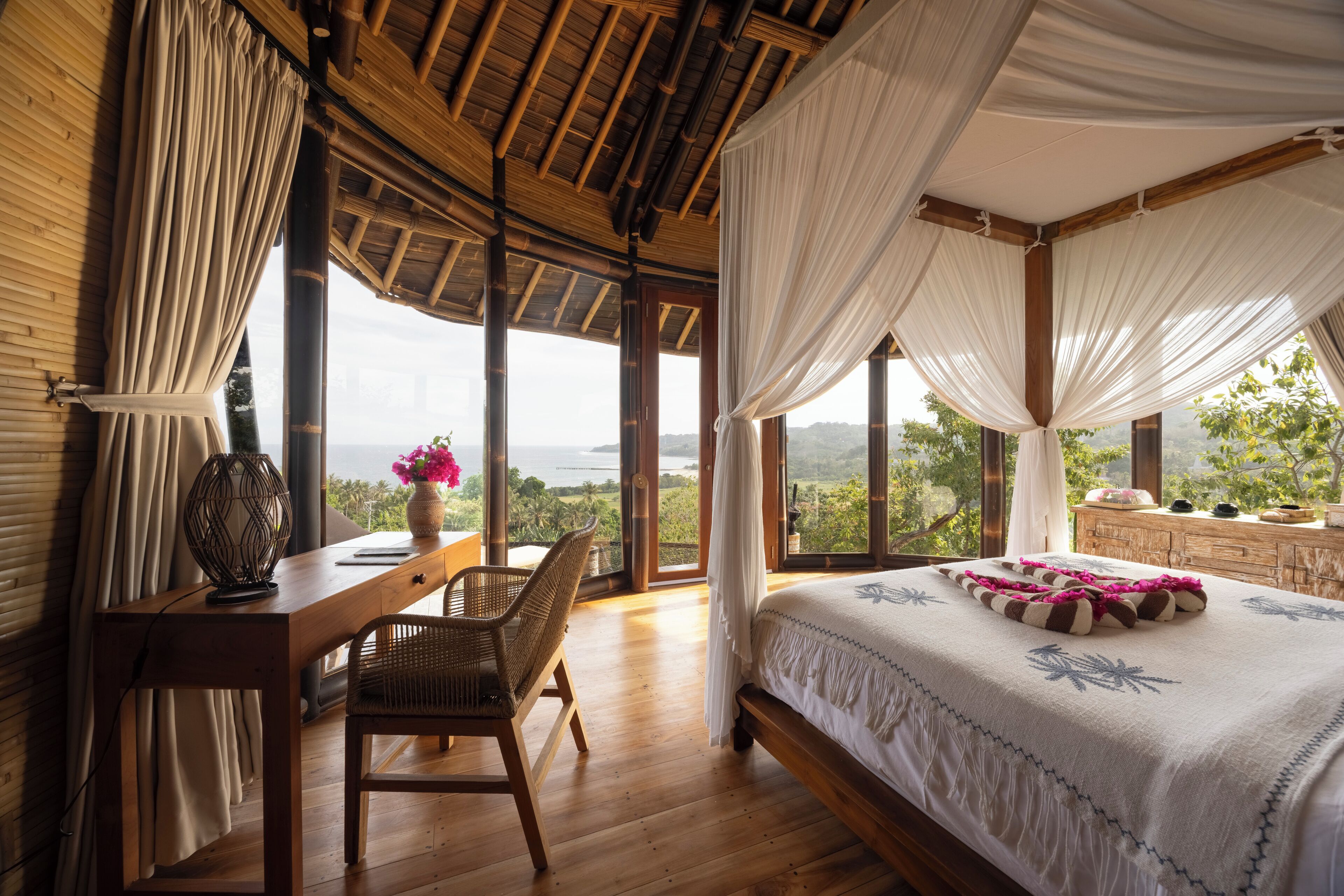 A bedroom with a wooden desk, a chair, a bed with a canopy, and large windows offering a view of the outdoors.