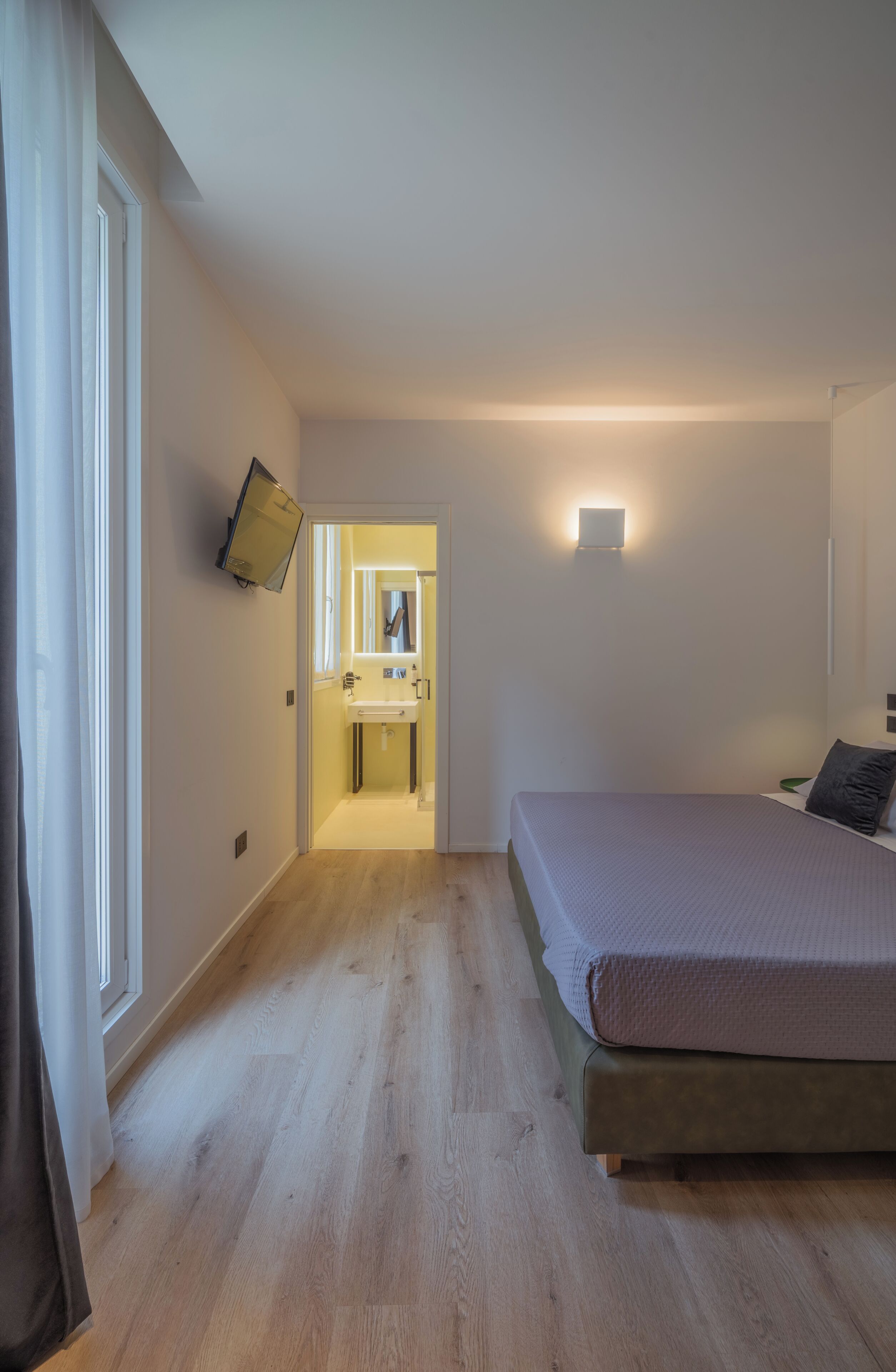A bedroom with a bed, a bedside table, a wall-mounted TV, and a bathroom visible through an open door.
