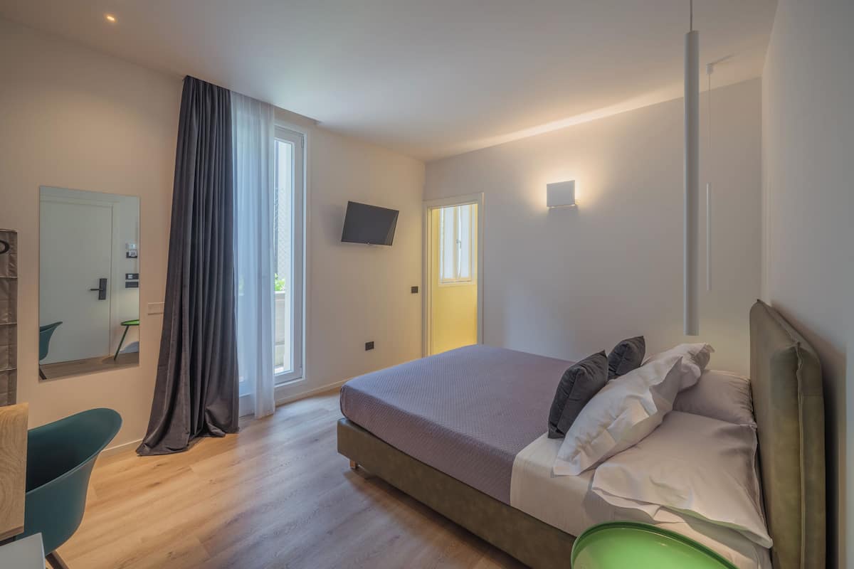 A modern hotel room with a large bed, a television, a mirror, and a window with curtains.