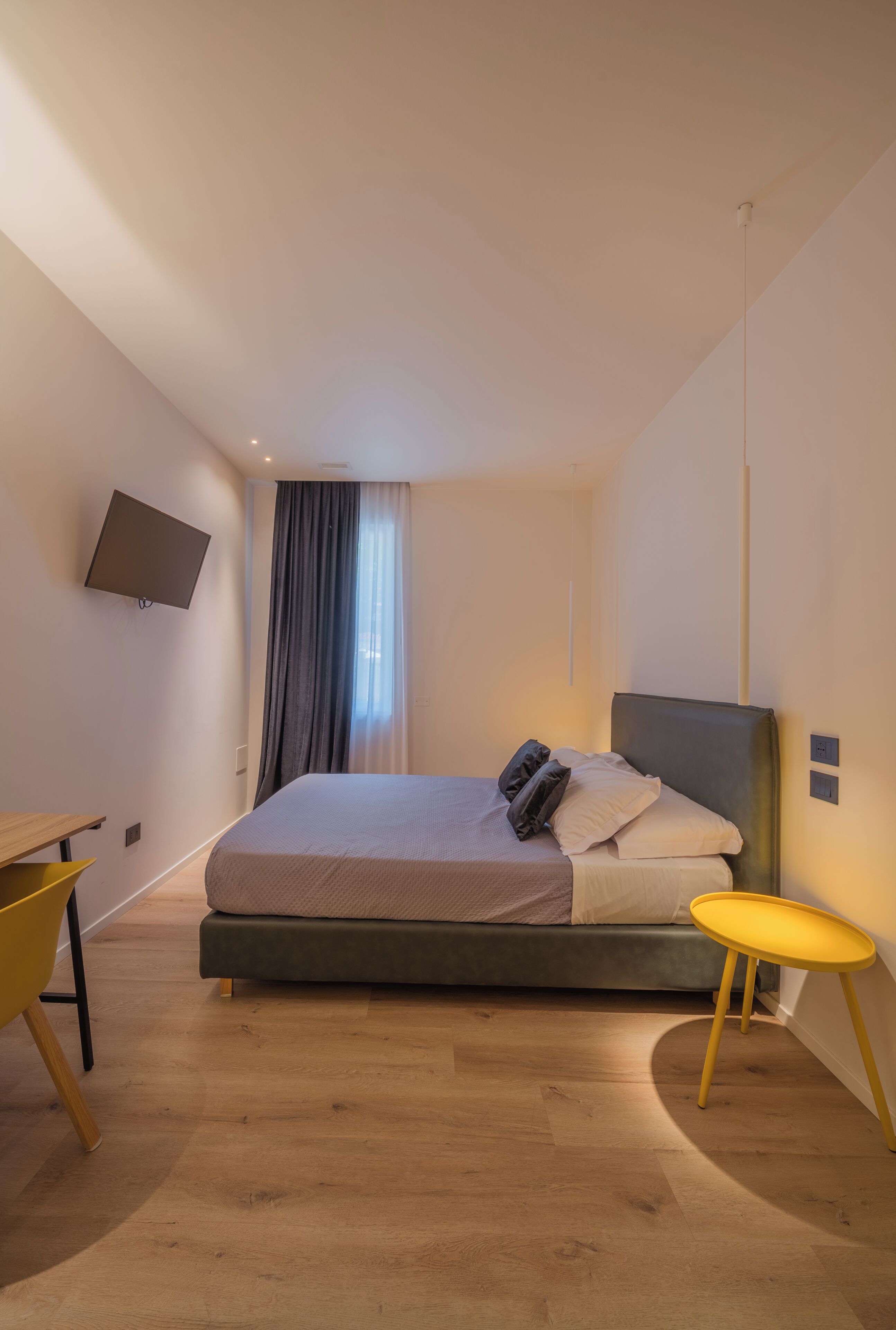 Um quarto moderno com uma cama, uma TV fixada na parede, uma mesinha e uma cortina.