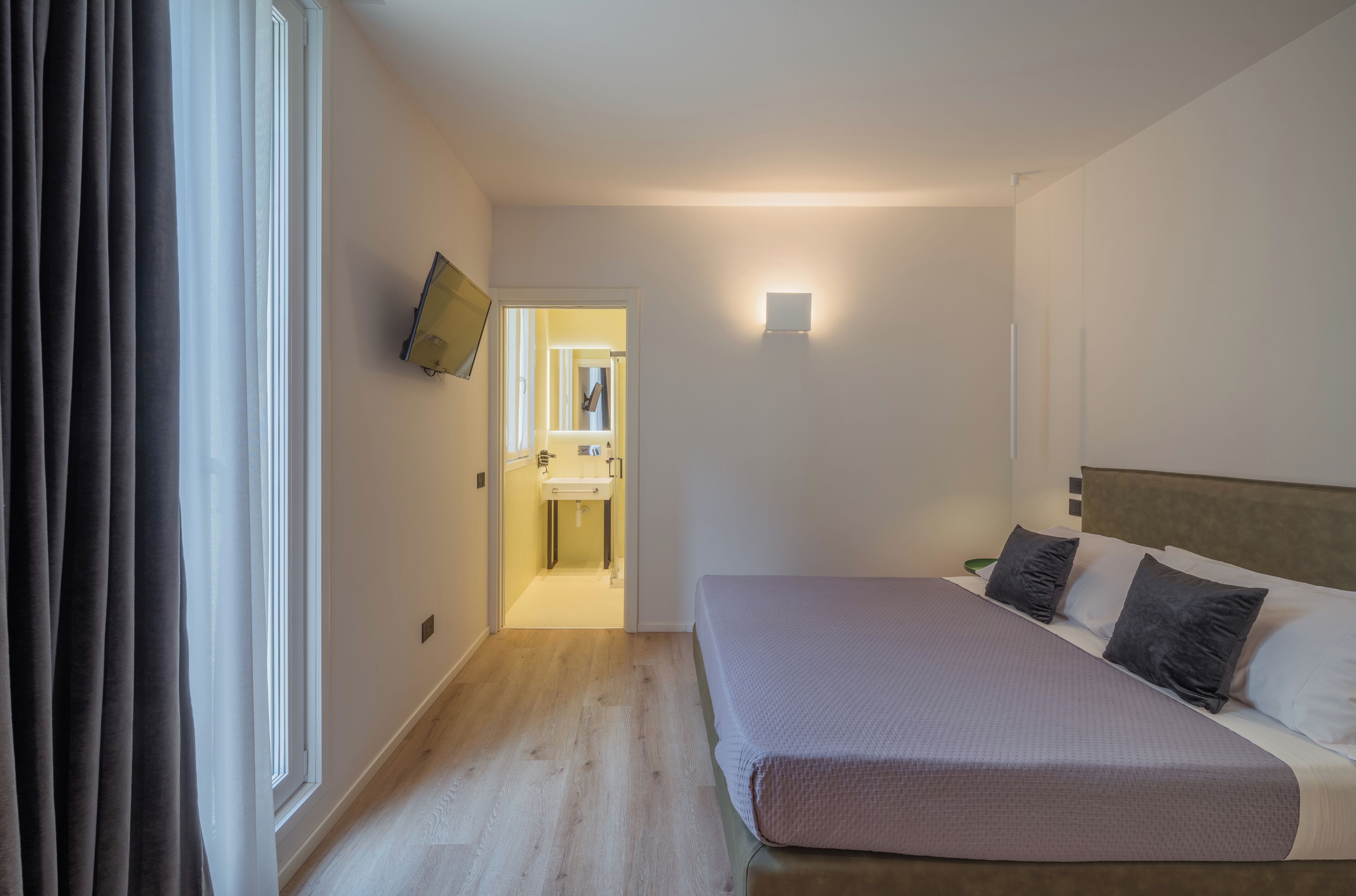 Habitación de hotel con cama, televisor de pantalla plana en la pared, mesita de noche y vista al baño.