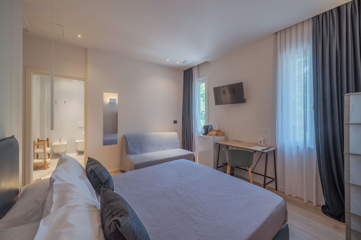 A hotel room with a bed, a desk, a chair, a TV, and a bathroom visible through an open door.