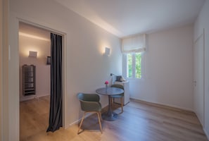 Ein Zimmer mit Esstisch, zwei Stühlen, einer kleinen Kommode, einem Fenster mit Vorhängen und einer Tür.