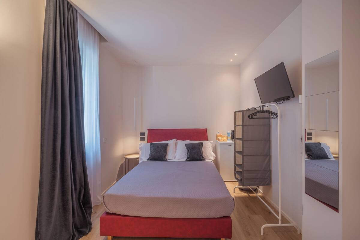 A hotel room with a bed, a TV, a bedside table, a wardrobe, and a window with curtains.