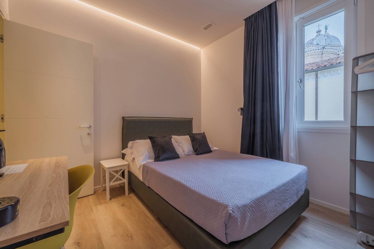 A modern hotel room with a bed, a desk, a chair, and a window with a view of a building.