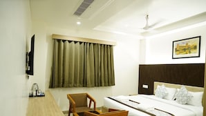 Une chambre d’hôtel avec un lit, un bureau et une chaise, une fenêtre avec des rideaux, un ventilateur de plafond et un tableau encadré au mur.