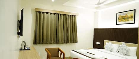 Une chambre d’hôtel avec un lit, un bureau et une chaise, une fenêtre avec des rideaux, un ventilateur de plafond et un tableau encadré au mur.