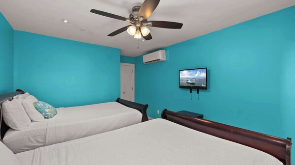 Una habitación con dos camas, ventilador de techo, televisor montado en la pared y aire acondicionado.