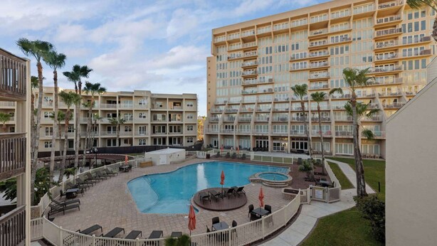 Ein Hotel mit einem zentral gelegenen Schwimmbecken, umgeben von Liegestühlen und Sonnenschirmen, mehreren Balkonen und Palmen.