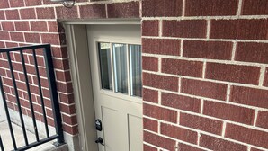 Fachada de ladrillo con puerta color beige, barandilla metálica y rejilla de metal en la base.
