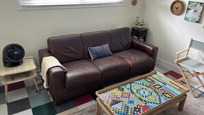 Un sofá de cuero marrón con un cojín, una mesita con un ventilador, una mesa de mosaico colorido, un sillón de director y una ventana con persianas.