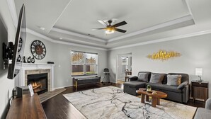 A modern living room with a fireplace, dark wooden flooring, a large rug, a gray sofa, and a ceiling fan. - The Naturehaus - Spacious 4-BR villa with gym/game room in fantastic Bentonville (Bentonville)
