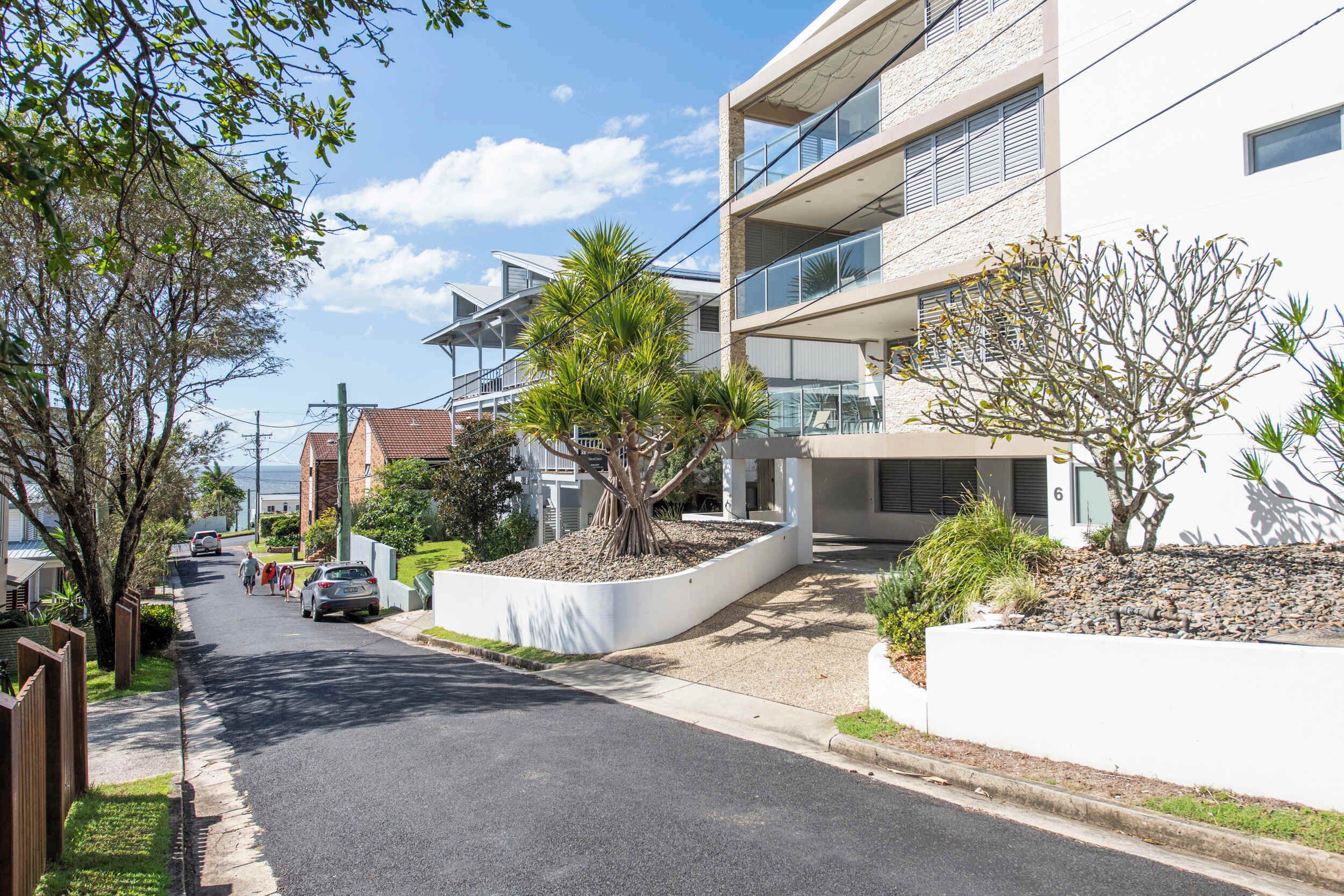 A modern residential street with a two-story house featuring a balcony, a driveway, and a well-maintained garden.
