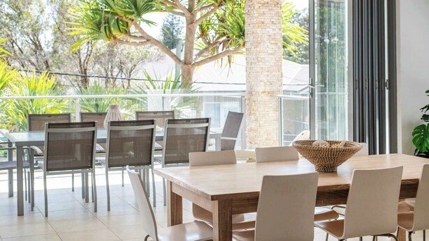 A modern dining area with a large table and chairs, a woven centerpiece, and a view of greenery outside. - Zinc One - Oceanstays - ocean views (Yamba)