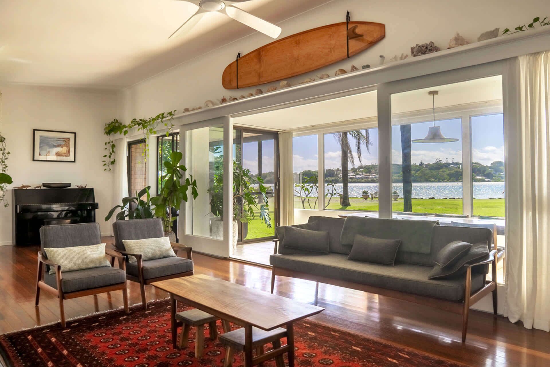 A spacious living room with a large window offering a view of a lake and greenery. There is a wooden coffee table, two armchairs, and a sofa.