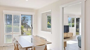 A spacious dining area with a large table and chairs, a TV in the background, and a sliding glass door leading to a garden. - Marg's - Oceanstays - Oceanfront (Brooms Head)