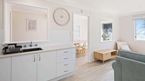 A modern kitchen with white cabinetry, a sink, and a clock on the wall. There is a dining area with a table and chairs, and a sofa in the living room. - Marg's - Oceanstays - Oceanfront (Brooms Head)