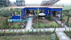 A blue building with a sign reading "The Tiger Waterfront." It has a patio area with tables and chairs, surrounded by plants and a pathway. - The Tiger Waterfront Resort (Puranpur)