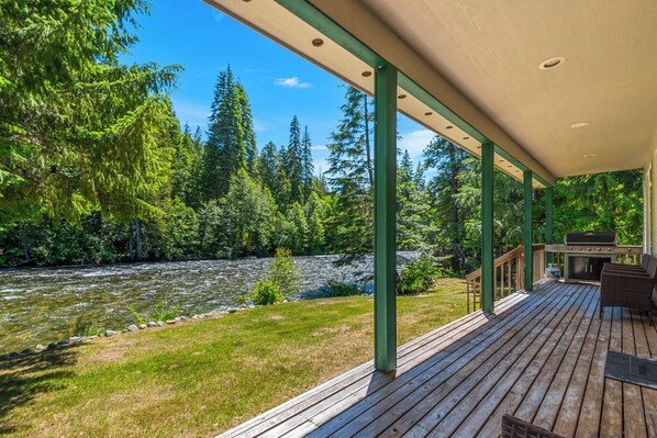 A wooden deck with a grill and seating area overlooking a river and forest. - Salmon's Rest - Rural Riverfront Retreat (Leavenworth)
