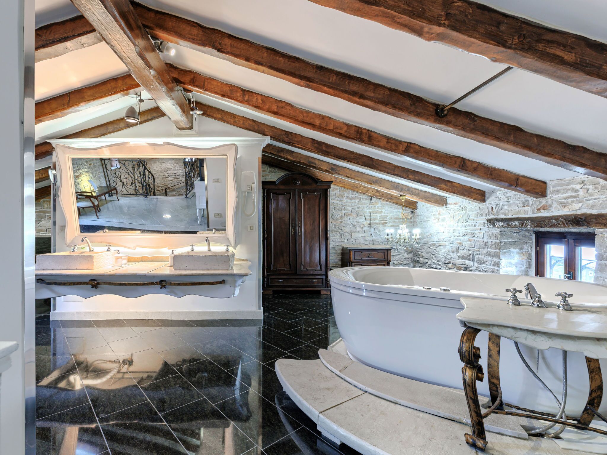 A modern bathroom with a large mirror, two sinks, a freestanding bathtub, and a stone wall.