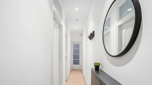 Um corredor com paredes brancas, soalho de madeira, um espelho redondo e uma pequena planta sobre um console escuro.