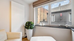 Un dormitorio moderno con un ventanal grande, una cama blanca, una silla blanca, una planta en maceta y un radiador.